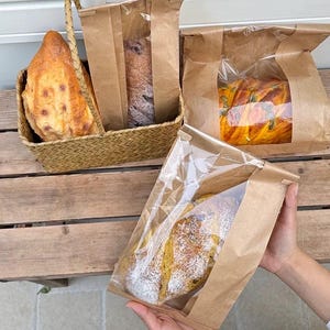 Op de afbeelding: Drie bruine papieren zakken met transparante plastic ramen, elk met een brood erin. Het brood is in verschillende vormen en kleuren, waaronder een rond brood met een spiraalpatroon, een lang brood met een knapperige korst en een kleiner brood met een lichte laag meel.