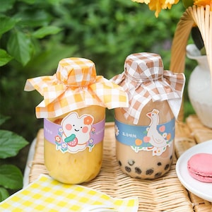 May include: Two glass bottles with colorful lids and labels. The bottles are filled with different beverages. One bottle has a yellow beverage and a cartoon character label. The other bottle has a brown beverage with a cartoon goose label. The bottles are on a wicker surface with a yellow and white checkered napkin.