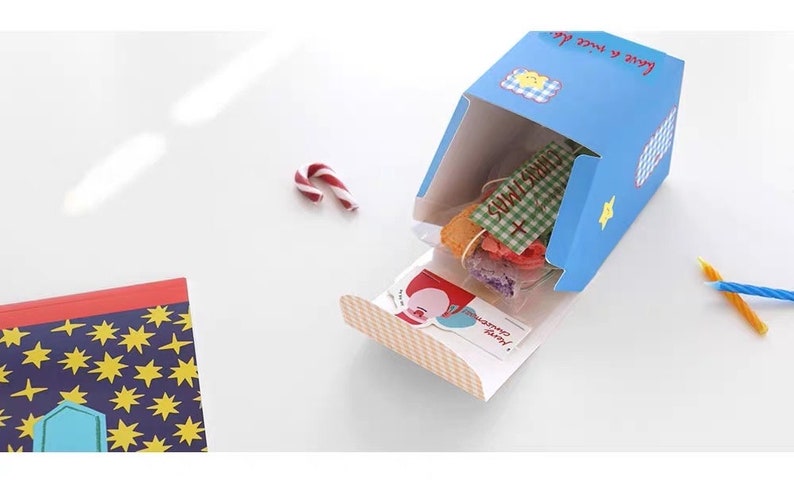 May include: A blue and white gift box with a checkered pattern and the text "Love & Sweet Life" on the side. The box is open and contains a variety of colorful candies and a small card with the text "Merry Christmas!"