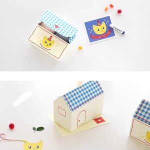 May include: A white and blue gingham patterned house-shaped gift box with a yellow cat face on the front. The box is open and the inside is lined with blue and white gingham paper. The box is sitting on a white surface. There is a small yellow cat face sticker on the front of the box.