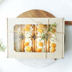 Puede incluir: Una caja de pasteles rellenos de mango y crema envueltos individualmente, atados con cuerda y una ramita de romero. La caja tiene una ventana transparente para ver los pasteles en el interior.