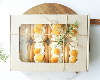 May include: A box of individually wrapped mango and cream filled pastries tied with twine and a sprig of rosemary. The box has a clear window to see the pastries inside.