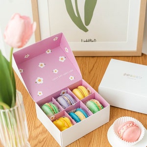 May include: A pink and white box with a floral pattern and the text "Lucky Day Lucky Life" contains colorful macarons. The box is sitting on a wooden surface next to a white box with the text "Sweet" and a pink macaron on a white plate.
