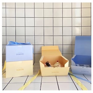 Puede incluir: Tres cajas de panadería, dos están apiladas una encima de la otra. La caja superior es azul claro con la palabra "BAKERY" impresa en ella. La caja inferior es amarilla con la palabra "BAKERY" impresa en ella. La tercera caja es amarilla con la palabra "BAKERY" impresa en ella y está abierta con un pastel dentro.