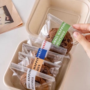 以下が含まれることがあります： 個包装されたチョコレートチップクッキーが入ったベージュ色の容器。各クッキーは、「SMALL HAPPINESS DELICIOUS」または「HAPPINESS」と書かれたカラフルなラベルが付いた透明なビニール袋に密封されています。
