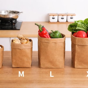 Könnte beinhalten: Vier braune Papiertüten in verschiedenen Größen, beschriftet mit S, M, L und XL, gefüllt mit Lebensmitteln. Die Tüten enthalten Eier, Ingwer, rote und grüne Paprika, Tomaten, Salat und Mais. Die Tüten stehen auf einer Holzoberfläche.