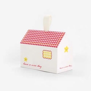 May include: A white gift box shaped like a house with a red and white checkered roof. The box has a yellow star with a smiley face on the side and the text "Have a nice day" on the front.