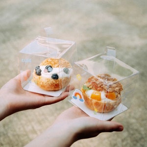 Peut inclure: Deux choux à la crème dans des boîtes en plastique transparentes. Un chou à la crème est garni de crème fouettée et de myrtilles. L'autre chou à la crème est garni de crème fouettée et d'une tranche d'orange. Les deux choux à la crème sont décorés d'autocollants colorés.