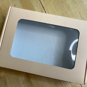 Puede incluir: Una caja de cart&oacute;n marr&oacute;n claro con una ventana transparente en la parte delantera. La caja est&aacute; vac&iacute;a.