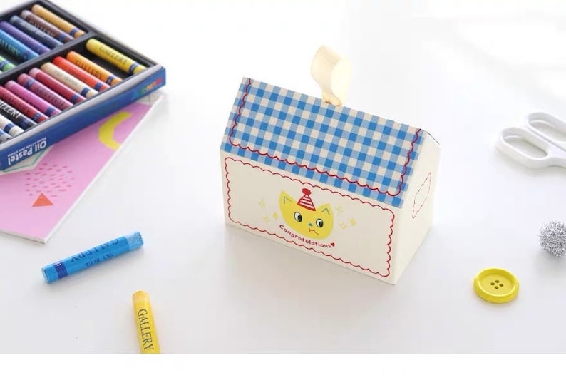 May include: A white house-shaped gift box with a blue and white checkered roof. The box has a yellow cat with a red party hat on the front and the text "Congratulations!" printed below the cat.