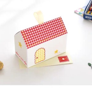 Könnte beinhalten: Eine kleine weiße Geschenkbox in Form eines Hauses mit einem rot-weiß karierten Dach. Die Box hat eine gelbe Tür und einen gelben Stern an der Seite. Der Text "have a nice day" ist an der Seite der Box aufgedruckt. Die Box steht auf einem gelben Umschlag mit einer roten Briefmarke.