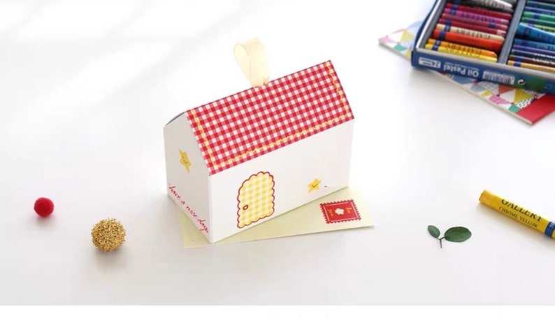 May include: A small white house-shaped gift box with a red and white checkered roof. The box has a yellow door and a yellow star on the side. The text "have a nice day" is printed on the side of the box. The box is sitting on a yellow envelope with a red stamp.