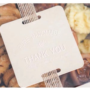 Puede incluir: Una etiqueta de regalo cuadrada blanca con el texto "PURE HANDMADE" y "THANK YOU" en relieve. La etiqueta est&aacute; atada con un lazo de cuerda marr&oacute;n y se coloca sobre una caja transparente llena de galletas surtidas.