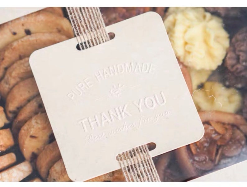 Puede incluir: Una etiqueta de regalo cuadrada blanca con el texto "PURE HANDMADE" y "THANK YOU" en relieve. La etiqueta est&aacute; atada con un lazo de cuerda marr&oacute;n y se coloca sobre una caja transparente llena de galletas surtidas.