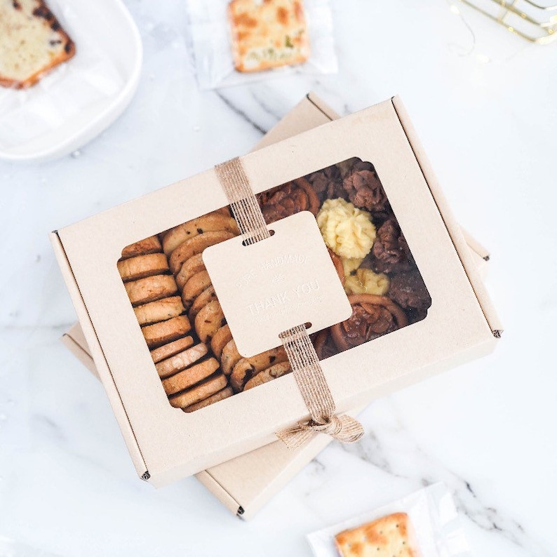 Puede incluir: Una caja de cart&oacute;n marr&oacute;n con una ventana transparente, llena de galletas surtidas. La caja est&aacute; atada con una cinta marr&oacute;n y tiene una peque&ntilde;a etiqueta que dice "Gracias".