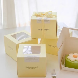 May include: Four yellow cardboard boxes with clear plastic windows, designed for packaging cakes. The boxes have the text "Delicious cake!" and "The best cake ever!" printed on them. One box contains a green cake with a white flower on top.