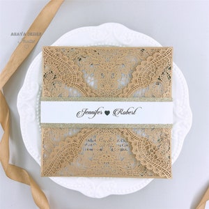 May include: A laser-cut wedding invitation with a delicate lace pattern in beige. The invitation features a white band with gold glitter and the names "Jennifer" and "Robert" with a heart symbol between them.
