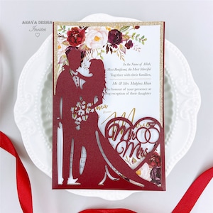 May include: Burgundy red wedding invitation with a laser cut design of a couple holding a bouquet of flowers. The invitation features a gold glitter border and a heart-shaped cutout with the words "Mr & Mrs" and two gold rings.