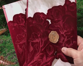 Könnte beinhalten: Zwei rechteckige Einladungskarten mit einem tiefroten Samteinband und einem komplexen geprägten Blumenmuster. Ein goldfarbenes Wachssiegel mit Rosenmotiv verschließt die Klappe. Die innere Karte ist weiß und bildet einen Kontrast zum roten Samt.