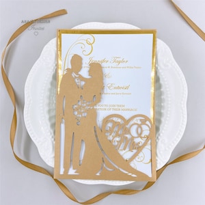 May include: A laser cut wedding invitation with a gold background and a brown silhouette of a bride and groom. The invitation features the text "Jennifer Taylor" and "Entwistl" with a heart shape and the words "Mr & Mrs" in the center.