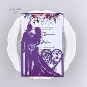 May include: Purple laser cut wedding invitation with a floral design and a silhouette of a couple. The text on the invitation reads "In the honour of your presence at the wedding of Leimaran to Naghavi, Friday, the twenty fourth of May, two thousand and twenty, at o'clock in the afternoon." The invitation also features a heart with the words "Mr & Mrs" and two rings.