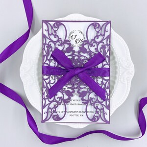 May include: Purple laser cut wedding invitation with a white background and a purple satin ribbon tied in a bow. The text on the invitation reads "Saturday August, Reception to start promptly at 7:00 PM, University of Washington Husky Union Building, Seattle, WA."