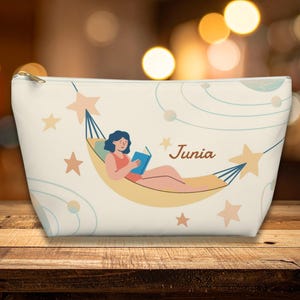 May include: A cream-coloured zippered pouch featuring a graphic illustration of a person with dark hair reading a blue book while resting in a yellow hammock. The design includes light orange stars, blue orbital lines, and the name Junia in brown script.