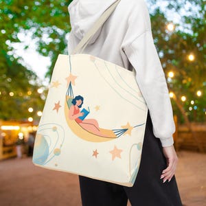 May include: A cream-coloured tote bag featuring a graphic illustration of a person with dark hair reading a blue book whilst resting in a yellow hammock. The design includes light orange stars, thin blue curved lines, and a blue planet on the side.