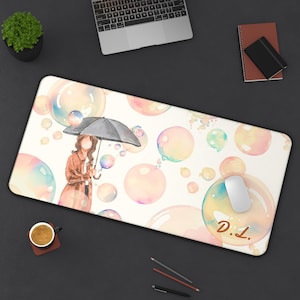 Può includere: Un tappetino da scrivania rettangolare con un'illustrazione ad acquerello di una persona con un cappotto marrone che tiene un ombrello nero tra numerose bolle colorate e traslucide. Il testo D.I. appare in marrone sul lato inferiore destro.