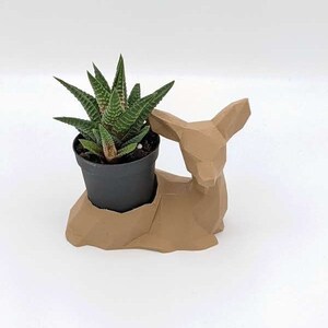 May include: A small, green succulent plant in a black pot, nestled in a geometric, tan-colored deer-shaped planter. The planter has a modern, angular design, creating a unique decorative piece for home decor.