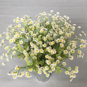 May include: A bouquet of white daisies with yellow centres arranged in a metal vase. The daisies are in full bloom and have a delicate, natural look.