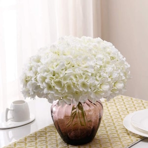 Pode incluir: Um buquê de flores de hortênsia brancas em um vaso de vidro transparente com um acabamento texturizado marrom-púrpura. O vaso está sobre um corredor de mesa com padrão amarelo e branco.