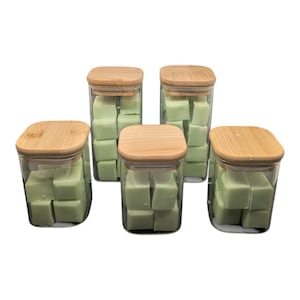 May include: Five clear glass jars with square wooden lids. Each jar is filled with light green, square-shaped wax melts. The jars are arranged in a slightly offset pattern against a white background.