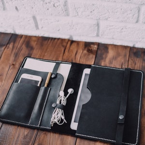May include: A black leather portfolio with white stitching detail. The portfolio has multiple pockets and compartments for holding a phone, pen, tablet, and headphones.