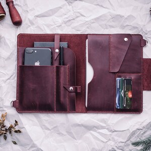 May include: A burgundy leather portfolio with multiple pockets and compartments. The portfolio is open and shows a smartphone, pens, and credit cards inside.