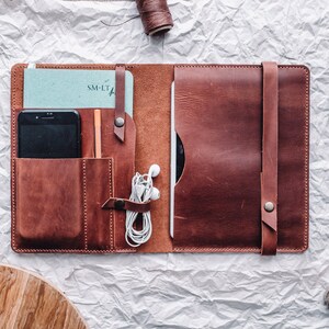May include: A brown leather portfolio with multiple pockets and a strap. The portfolio is open to reveal a notebook, a phone, a pencil, and headphones.