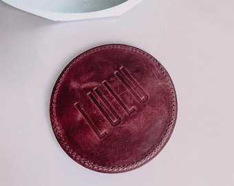 May include: A round, burgundy leather coaster with the word "LULU" embossed in the center. The coaster has a stitched edge and is set against a light background. A light blue, geometric-shaped object is in the background.