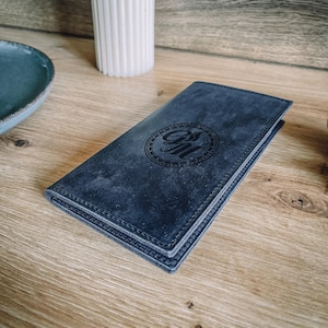 Op de afbeelding: Een marineblauwe leren portemonnee met een cirkelvormig reliëf ontwerp. De portemonnee is rechthoekig en lijkt gesloten. De achtergrond omvat een witte kaars en een blauwe kom op een houten oppervlak.