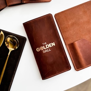 May include: Several brown leather menu holders are displayed, each embossed with "THE GOLDEN BALL" in gold. One is open, showing a pocket. A gold spoon and fork sit on a black leather placemat.