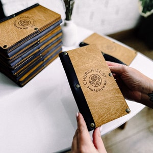 Puede incluir: Una pila de cubiertas de menú de madera con una encuadernación de cuero negro y el texto "Churchill St. Shoreview, MN" grabado en la parte delantera. La cubierta del menú se sostiene en una mano.