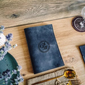 Może przedstawiać: Granatowa skórzana okładka menu z okrągłym monogramem. Złota łyżka i widelec spoczywają na lnianej serwetce. Widoczny jest również kieliszek do wina i podkładka.