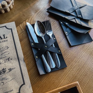 May include: A black leather napkin holder with a silver fork, knife, and spoon wrapped inside. The holder has decorative brass rivets.