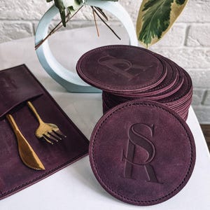 May include: A set of six burgundy leather coasters with a monogrammed initial "S" in a decorative font. The coasters are stacked on top of each other and are sitting on a white surface.