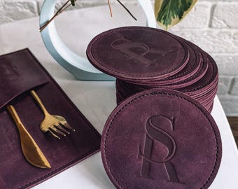 May include: A set of six burgundy leather coasters with a monogrammed initial "S" in a decorative font. The coasters are stacked on top of each other and are sitting on a white surface.
