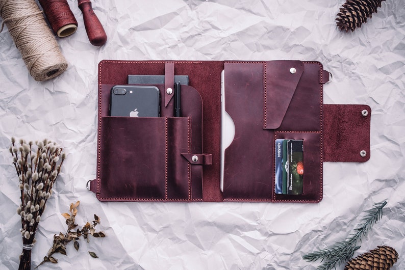 May include: A burgundy leather portfolio with multiple pockets and compartments. The portfolio is open and shows a smartphone, pens, and credit cards inside.
