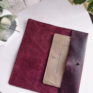 May include: A burgundy leather folder with a brown paper insert and a flap closure. The folder has a small brass button on the flap.