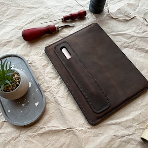 May include: A brown leather tablet sleeve with a flap and a pen holder. The sleeve is on a crumpled brown paper background.