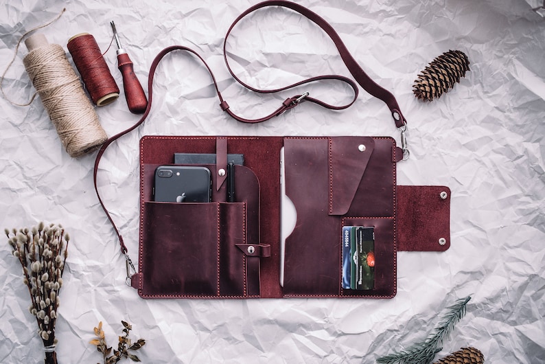 May include: A burgundy leather portfolio with multiple pockets and a strap. The portfolio is open and shows a phone, pens, and credit cards inside.