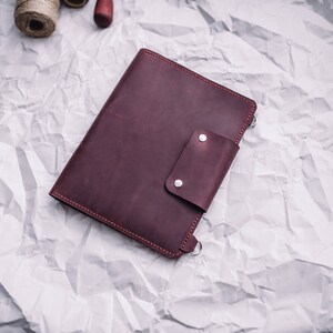 May include: A burgundy leather notebook with a flap closure and a silver snap. The notebook is on a white crumpled paper background.