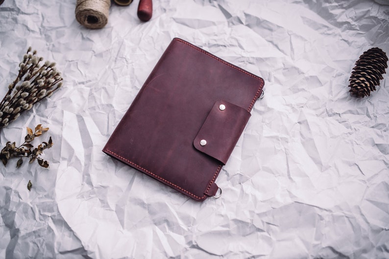 May include: A burgundy leather notebook with a flap closure and a silver snap. The notebook is on a white crumpled paper background.